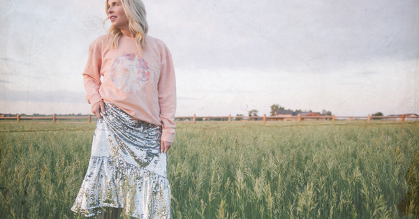 Woman wearing a pink graphic sweatshirt and silver sequin skirt standing in a grassy field at sunset — Miracle Magnet Co. inspirational apparel for women, collage-style design, soft crewneck sweatshirt.